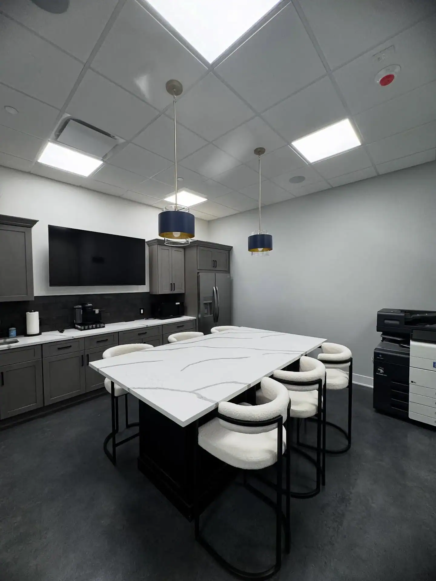 Modern office kitchen featuring a marble island, six chairs, TV, cabinets, and epoxy flooring.