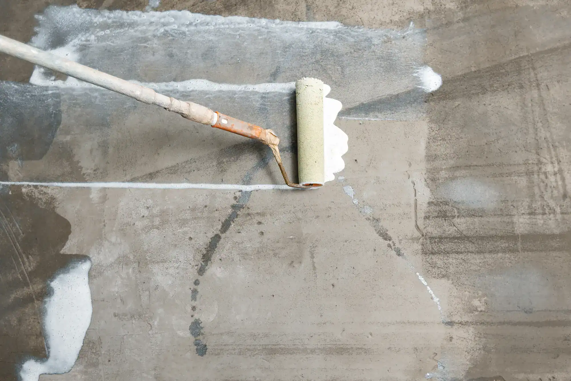 A paint roller applies white paint to a rough gray wall section by an Epoxy Flooring Contractor in Bohemia, NY.